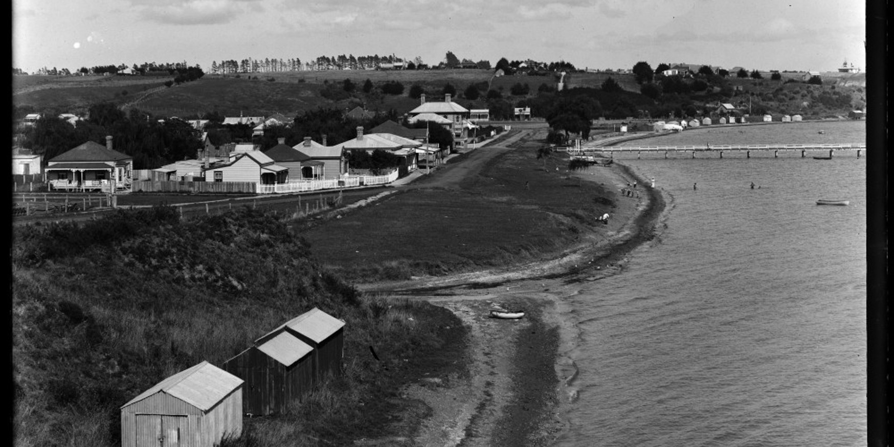 Looking west along St Heliers Bay Beach, showing St Heliers Bay Wharf, boatsheds, the intersection of Cliff Road and Tamaki Drive and St Heliers Bay Hotel